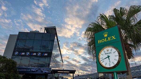 Exterior view of a modern glass building with BMW and Rolex branding at a racetrack, featuring a large Rolex clock and palm trees under a colorful evening sky.