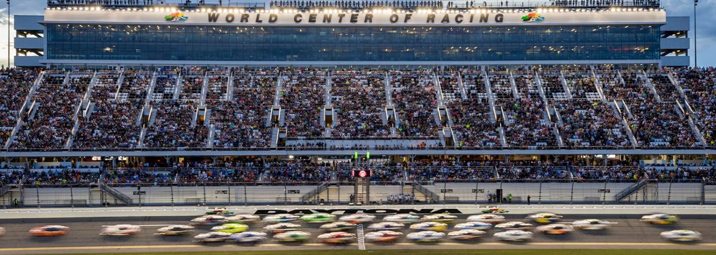 Blurred NASCAR race cars speed past the start-finish line at Daytona International Speedway as a packed grandstand watches under the “World Center of Racing” sign.