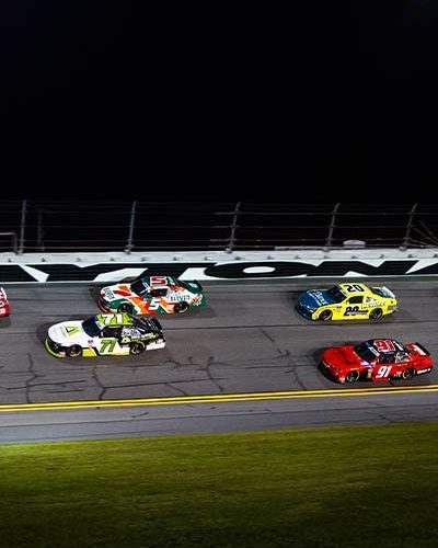 A group of NASCAR race cars speed side-by-side on the track at night at Daytona International Speedway. The cars are painted in various bright colors, including red, green, yellow, and orange, with sponsor logos visible. The word “DAYTONA” is painted in large white letters on the outer wall behind them under bright track lights.