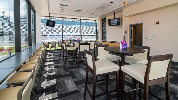 An indoor premium suite at Daytona International Speedway with large windows overlooking the track. The room features high-top tables and cushioned bar chairs arranged neatly along the window and throughout the space. Two wall-mounted TVs are visible, and the view outside shows the grandstands with the text “World Center of Racing.” Natural light fills the room, highlighting the modern, clean design.
