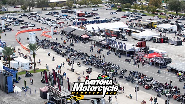 Aerial view of the Daytona Motorcycle Marketplace event at Daytona International Speedway, showing rows of motorcycles parked along wide walkways filled with visitors. Numerous vendor tents and trailers line the area, displaying banners and merchandise, while trucks and portable restrooms are visible in the background. The foreground features palm trees, signage, and a “Daytona Motorcycle Marketplace” logo in the bottom left corner.