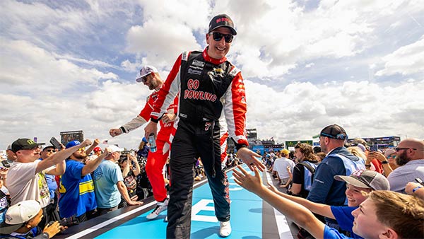 A NASCAR driver wearing a black and red ‘Go Bowling’ firesuit walks along a fan walkway, smiling and reaching out to high-five excited fans lined along the barrier. Another driver in a red suit follows behind. The scene is bright and energetic under a partly cloudy sky, with a large crowd gathered around the walkway.