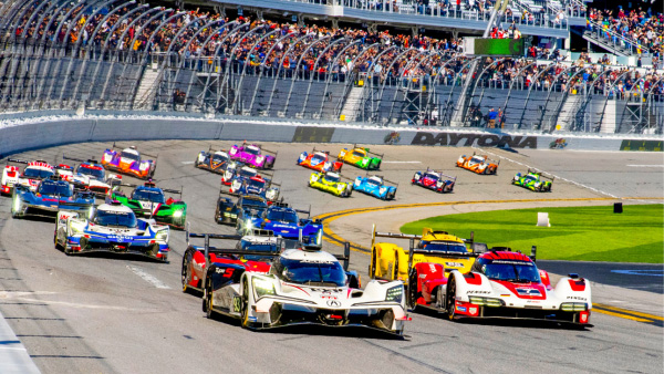 A large pack of IMSA prototype and GT race cars speeds through the Daytona International Speedway tri-oval, led by white and red prototypes, with colorful cars close behind, grandstands full of spectators on the left, and the green infield grass on the right under bright daylight.