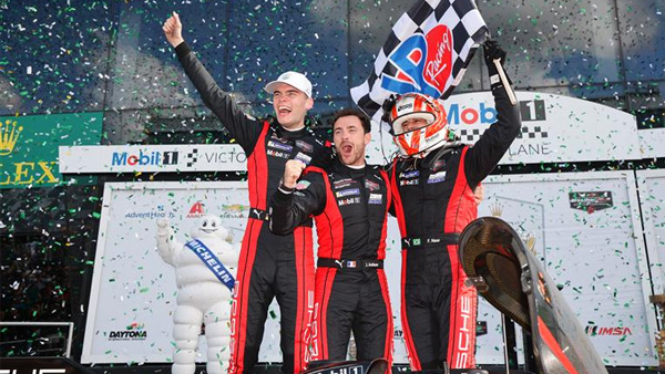 Three race car drivers in matching black and red racing suits celebrate on a podium, raising their arms as green confetti falls. One driver holds a checkered flag. A Michelin Man figure and Daytona International Speedway branding appear in the background.”