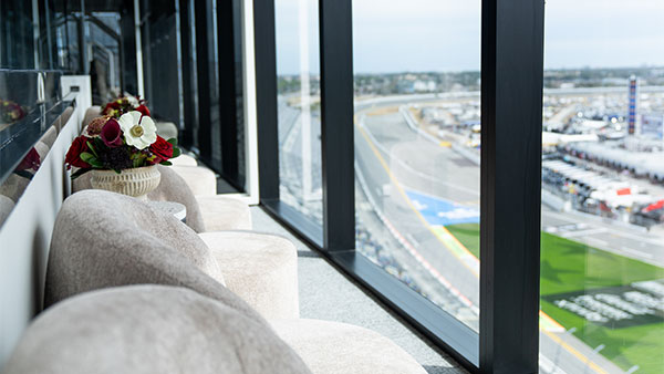 Luxury suite seating area at Daytona International Speedway overlooking the track through floor-to-ceiling windows. A light-colored upholstered couch sits beside a small table with a floral arrangement, while the racetrack, infield grass, and pit road are visible outside below.