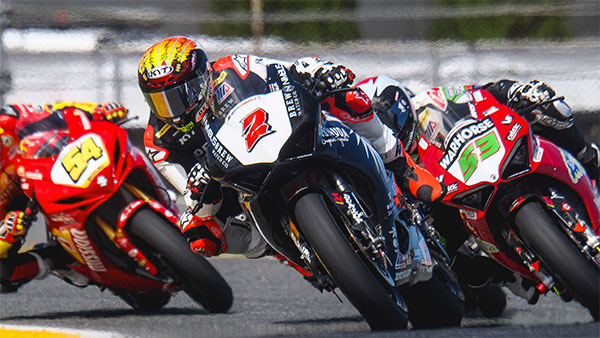 Three sport motorcycles lean sharply into a turn during a road race, with riders in full racing leathers and helmets battling closely for position on the track.