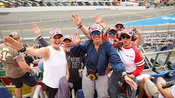 A group of fans wearing racing-themed hats and casual clothing stand in stadium seats, smiling and waving toward the camera. The racetrack and catch fence are visible behind them as they pose enthusiastically during a race event.