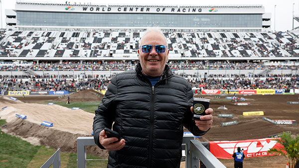 Man wearing sunglasses and a black jacket stands in front of a dirt race track at Daytona International Speedway, holding a drink and a phone, with grandstands and spectators in the background.”