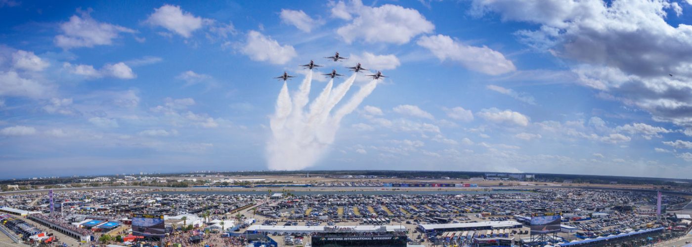 A wide aerial view of Daytona International Speedway shows a large crowd and expansive parking areas filled with vehicles and event tents. Above the venue, a group of jets flies in a tight formation, releasing white smoke trails that fan outward in the sky. The sky is bright blue with scattered clouds, creating a dramatic backdrop for the flyover. Below, the speedway grounds are busy with spectators, structures, and activity areas, conveying the scale and energy of a major race day event.