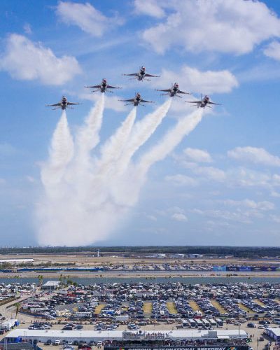 A wide aerial view of Daytona International Speedway shows a large crowd and expansive parking areas filled with vehicles and event tents. Above the venue, a group of jets flies in a tight formation, releasing white smoke trails that fan outward in the sky. The sky is bright blue with scattered clouds, creating a dramatic backdrop for the flyover. Below, the speedway grounds are busy with spectators, structures, and activity areas, conveying the scale and energy of a major race day event.