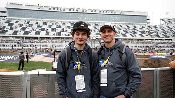 The image shows two people standing side by side at track level inside Daytona International Speedway. Both are wearing dark hooded sweatshirts, baseball caps, backpacks, and event credentials on lanyards around their necks. They are smiling and facing the camera. Behind them is a dirt racing track set up on the infield, with sculpted jumps and turns visible. A barrier separates them from the track area. In the background, the large grandstands rise up, partially filled with spectators. At the top of the grandstands, the “World Center of Racing” signage is visible on the main building facade.