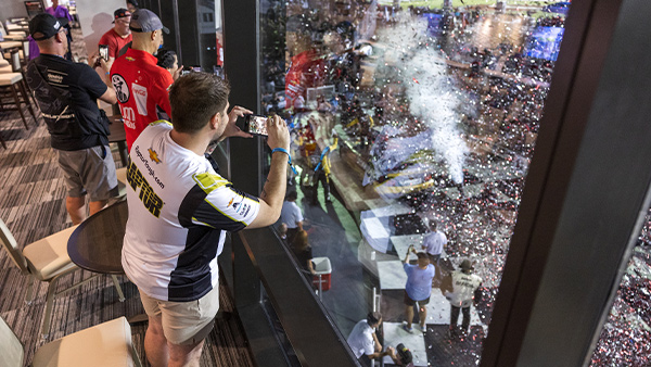 Guests inside a premium hospitality suite stand along floor-to-ceiling windows overlooking a race celebration below. One guest in the foreground photographs the scene with a smartphone as confetti fills the air outside. Additional guests watch from the suite while team members and event staff gather near the celebration area beneath the windows.