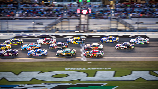 A pack of NASCAR stock cars races side-by-side across the start/finish line at Daytona International Speedway during a night event. Multiple brightly colored cars run three- and four-wide in close competition, with grandstands full of spectators visible in the background. The painted “DAYTONA” lettering and start/finish stripe are visible on the track surface in the foreground.