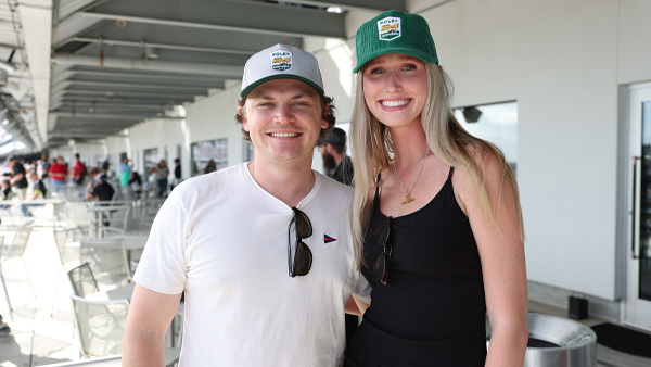 Two guests stand together on an outdoor hospitality terrace overlooking a venue concourse. Both are smiling at the camera and wearing green-and-white event-branded caps. The person on the left wears a white T-shirt with sunglasses hanging from the collar, while the person on the right wears a black sleeveless top with sunglasses hanging at the neckline. Tables, chairs, and other guests are visible in the background beneath a covered structure.