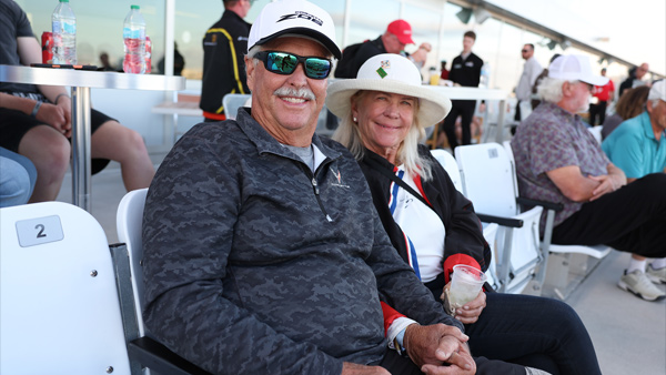 Two guests sit together in reserved grandstand-style seating within a hospitality area. The person on the left wears a dark patterned quarter-zip pullover, sunglasses, and a white cap, while the person on the right wears a light-colored wide-brim hat, dark jacket, and white shirt. Both are smiling toward the camera. Additional guests, tables, and refreshments are visible in the background.