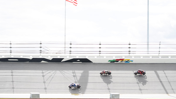 Three motorcycle racers compete on the high banks of Daytona International Speedway, with the DAYTONA logo and an American flag visible in the background.