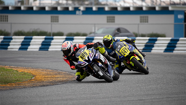 Two motorcycle racers lean into a turn while competing closely on the Daytona International Speedway road course.